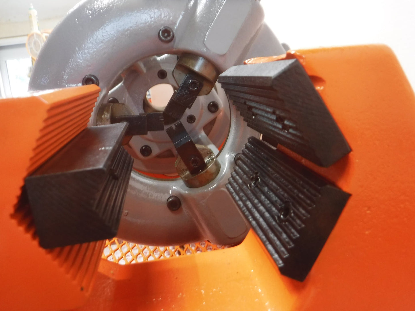 Close-up of the jaws on an Aeroquip FT1013-1-5 Reusable Hose Assembly Machine (Reconditioned), showing a quick acting wrench-type chuck with three black metal clamps and orange housing. Blurry background.