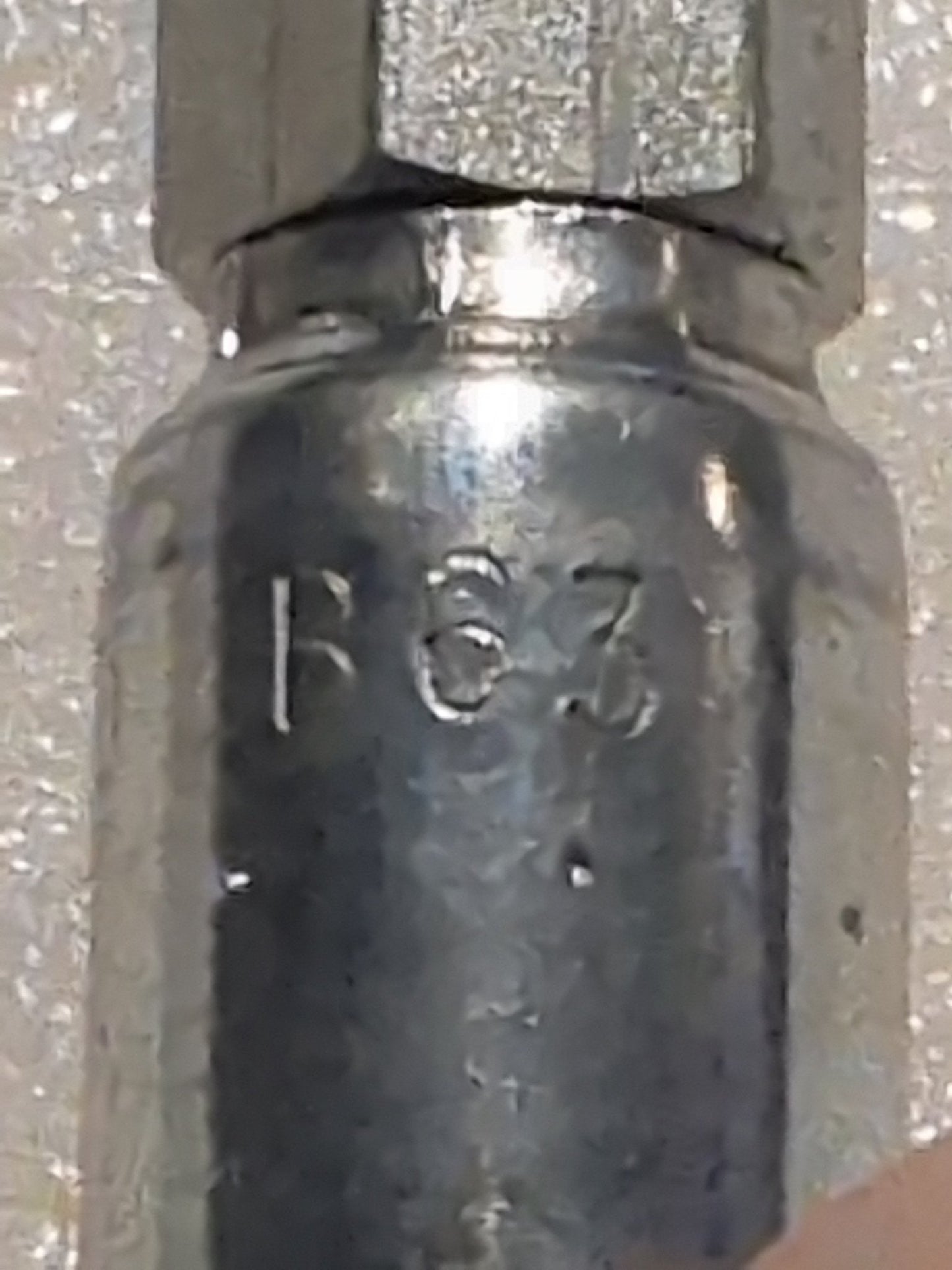 Close-up of a 04U-B63 Weatherhead 1/4 hose x 3/16 inverted male fitting (CS20-4X3) with “B63” engraved on its metal surface. A light, speckled background and part of a hand or finger are also visible.