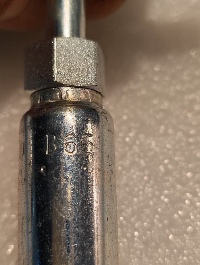 Close-up of a Weatherhead 04U-B65 (1/4 Hose x 5/16 Inverted Male Fitting, CS20-4X5) metallic fitting with B65 engraved, commonly used in hydraulic hose assemblies. The background is textured and light-colored.
