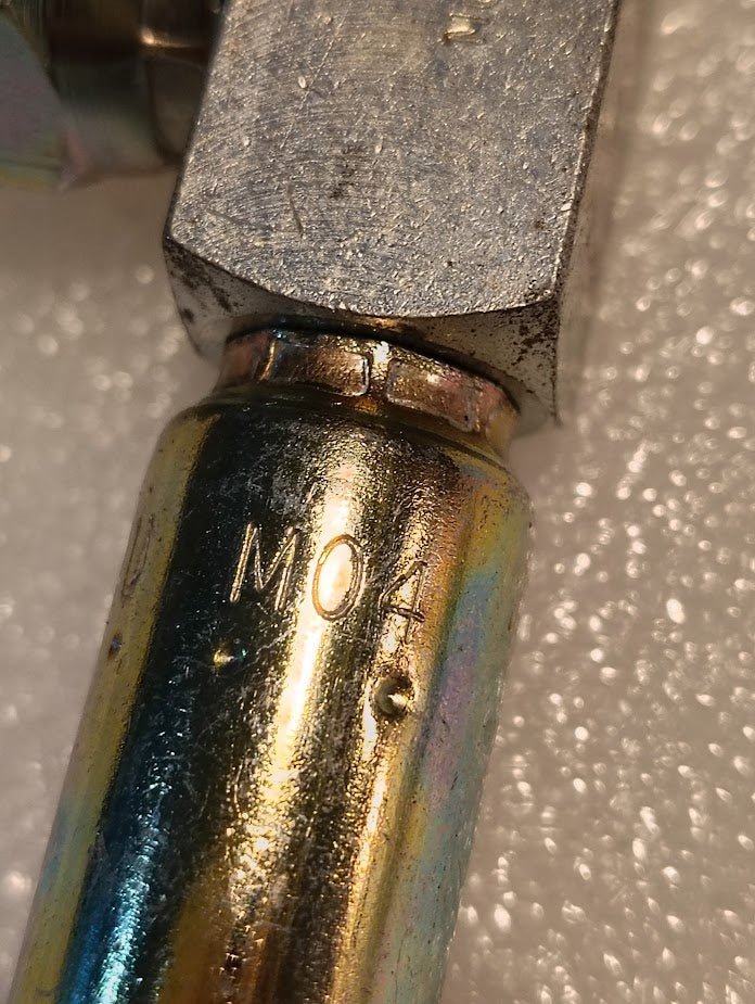 Close-up of a Weatherhead 04U-M04 1/4 hose x 1/4 NPTF male fitting (CS14-4X4) with M04 engraving, resting on a textured white surface. Slight discoloration is visible; commonly used in 100R style hoses.