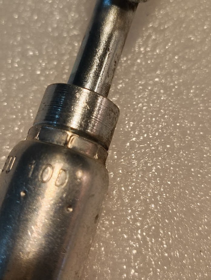 Close-up of a shiny metallic 06U-10D Weatherhead 3/8 Hose x M16x1.5 DIN Female Fitting (CS41-6X10) with engraved numbers, including 10D, lying on a textured light surface.