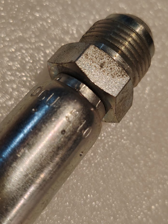 Close-up of a Weatherhead 06U-510 (3/8 Hose x 5/8 JIC Male Fitting, CS08-6X10) metal pipe fitting with hex nut and threaded end, used in hydraulics, resting on a textured light surface with faint markings visible.