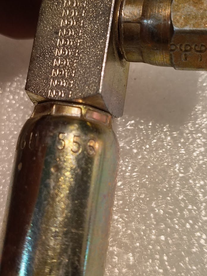 Close-up of a 06U-556 Weatherhead 3/8 Hose x 3/8 JIC Female Fitting (CS17-6X6) attached to a cylindrical canister, embossed numbers and letters visible, set against a textured white background.