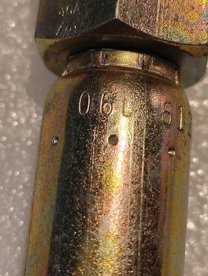 Close-up of a Weatherhead 06U-612 (3/8 Hose x 3/4 JIC Female Fitting, CS09-6X12) metallic cylinder, engraved with 06761 and two raised dots. The worn surface has a reflective, multicolored finish and textured background.