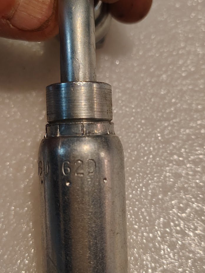 A person’s fingers hold a Weatherhead 06U-62D (3/8 Hose x M18x1.5 DIN Female Fitting, CS44-6X12) with “62D” embossed, set against a textured white background.