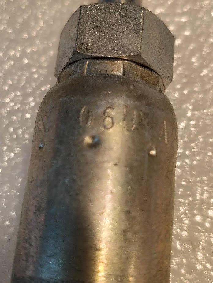 The image shows a close-up of an aged metallic cylindrical object featuring a hexagonal Weatherhead 06U-A24 fitting at the top. The surface has markings, including 06UXA, and the textured background resembles bubble wrap.