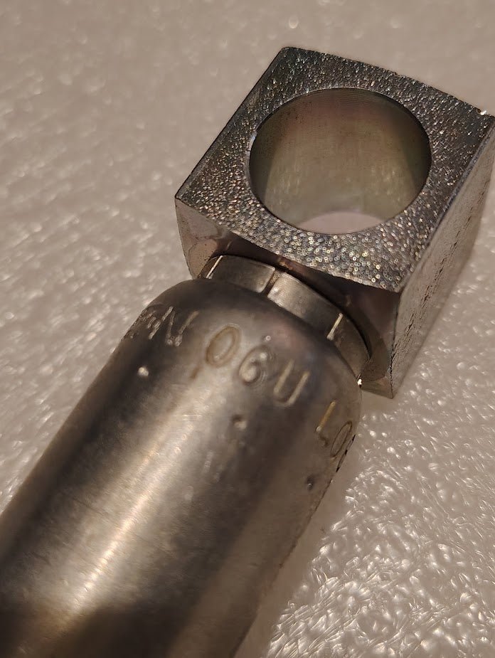 Close-up of a Weatherhead 06U-L06 3/8 Hose x 3/8 Banjo Fitting - 90 bend, attached to a square metal block with a circular hole, on a textured white surface. Partially visible engraved letters and numbers appear on the fitting.