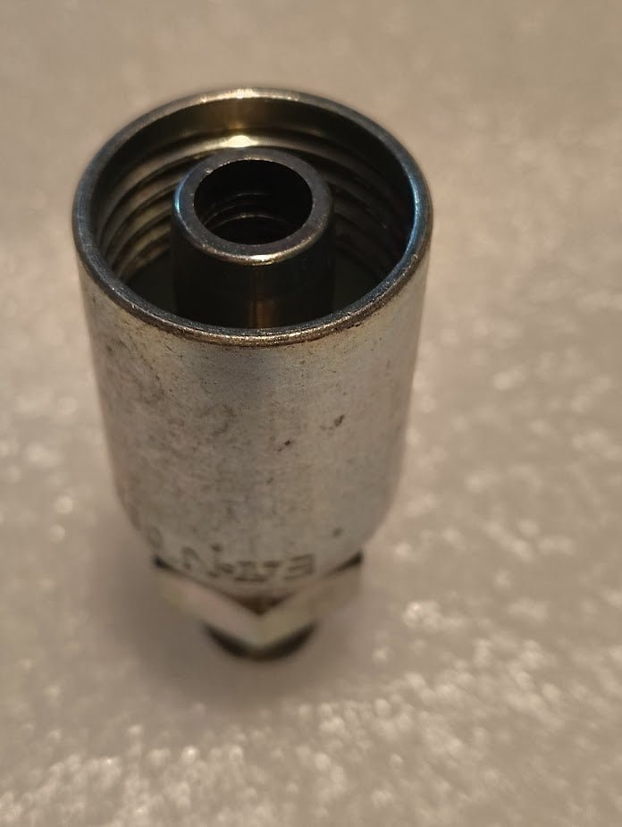 A close-up of the 08U-104 Weatherhead 1/2 Hose x 1/4 NPTF Male Fitting (CS01-8X4), a metal hydraulic hose fitting with internal threading, shown upright on a textured, light-colored surface.