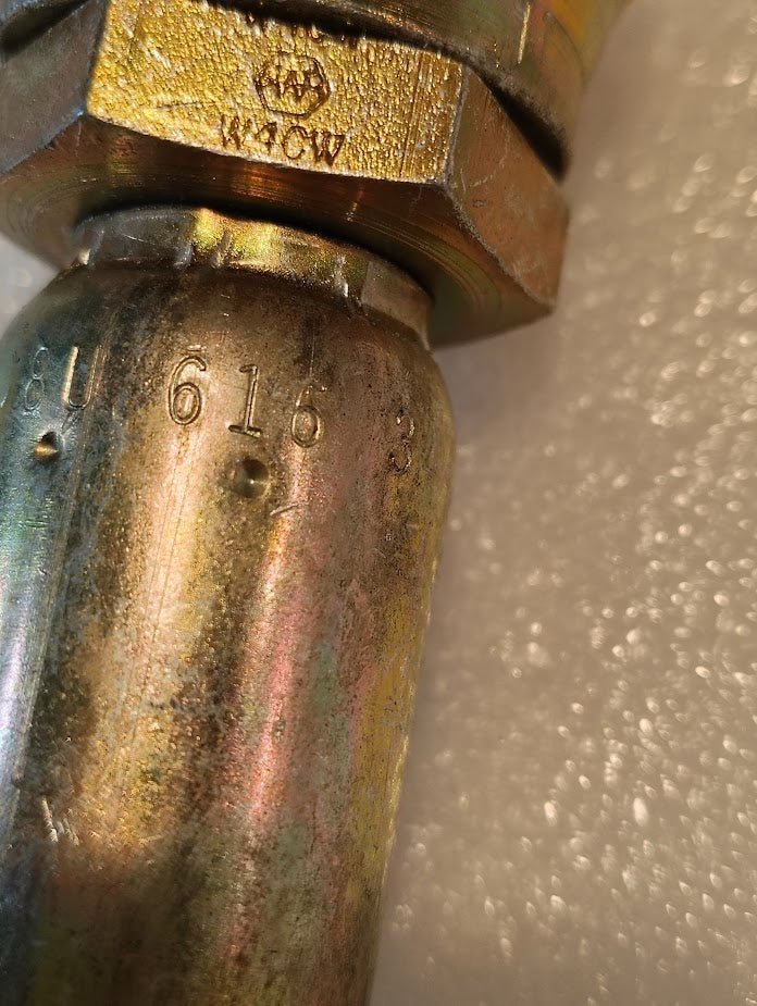 A close-up of a metallic 08U-616 Weatherhead fitting shows a hexagonal nut and brass-colored surface with engraved markings. The slight iridescent sheen contrasts against a textured white background, highlighting its intricate details.