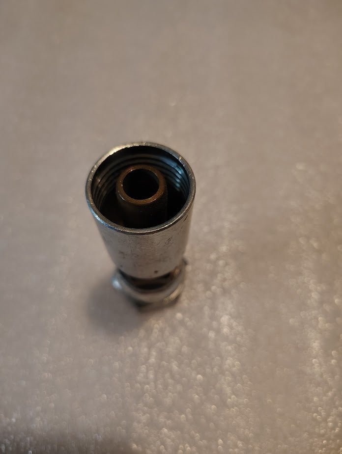A close-up of a metallic cylindrical object shows an 08U-66C Weatherhead 1/2 Hose x M24x1.5 DIN Female Fitting with a threaded opening, placed on a textured gray surface.