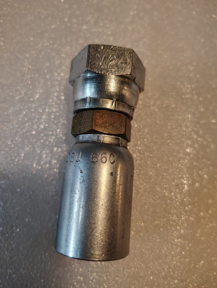 A close-up of the 08U-66C Weatherhead 1/2 Hose x M24x1.5 DIN Female Fitting (CS38-8X16), showing hexagonal and cylindrical sections with markings, rests on a textured white surface.