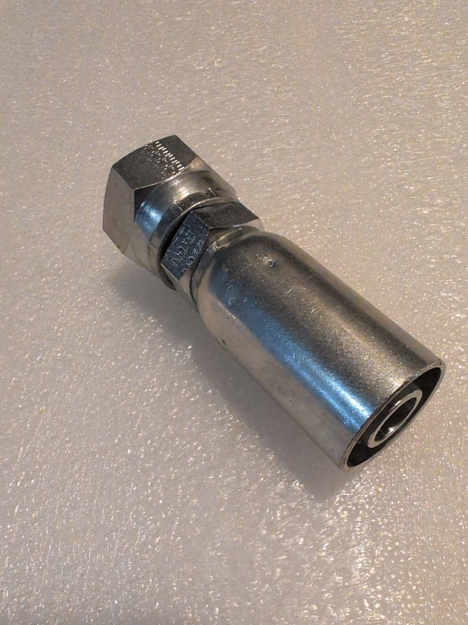 A close-up of the Weatherhead 10U-612 (5/8 Hose x 3/4 JIC Female Fitting, CS09-10X12), a metal hydraulic fitting with hex nut and cylindrical body for 100R1 hoses, on a textured light surface.