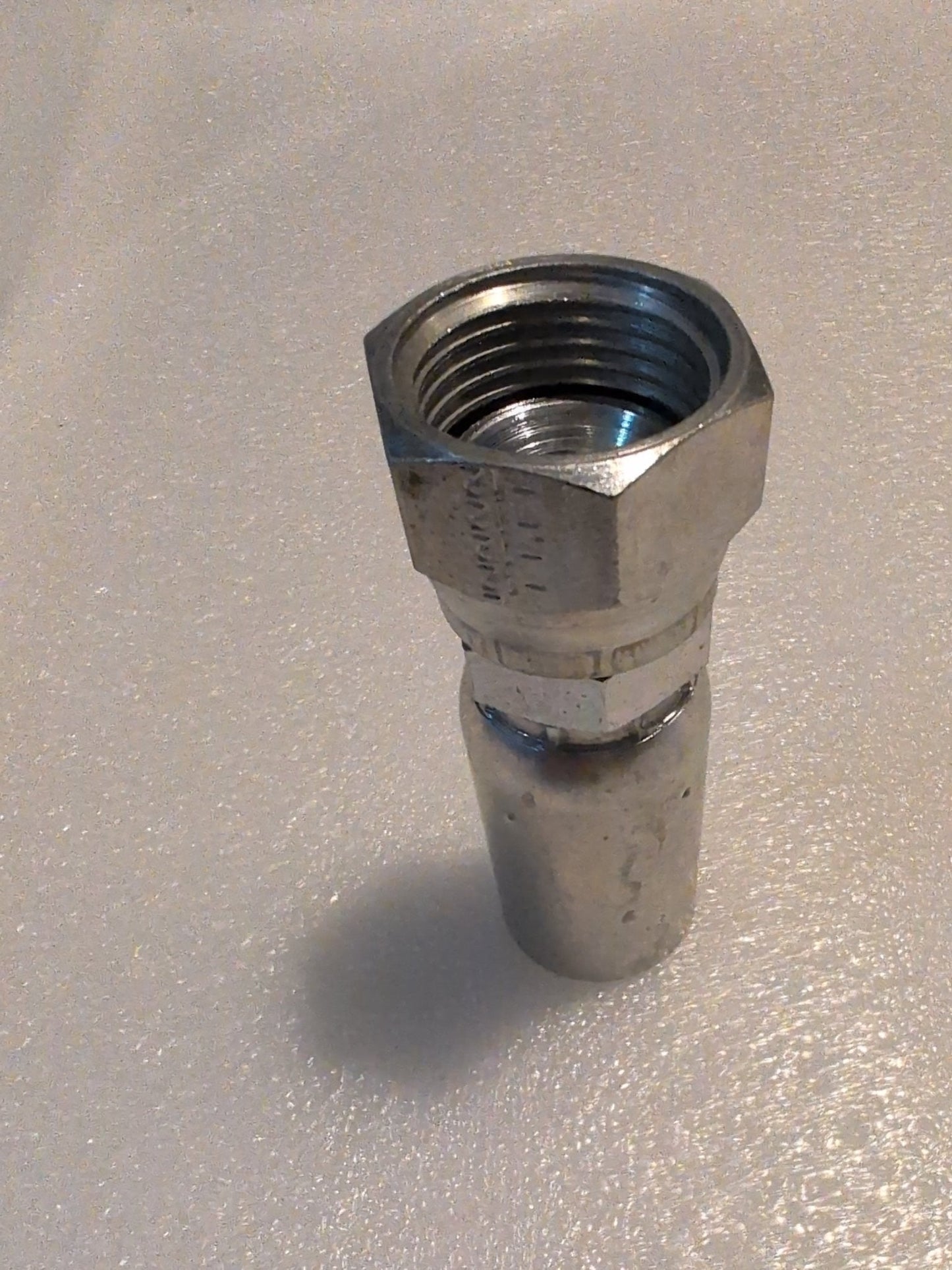 The 10U-612 Weatherhead fitting, featuring a metallic hexagonal nut with internal threading, is compatible with 100R1 hydraulic hose. Its displayed upright on a white textured surface, likely for plumbing or mechanical use, with shadows to the left.
