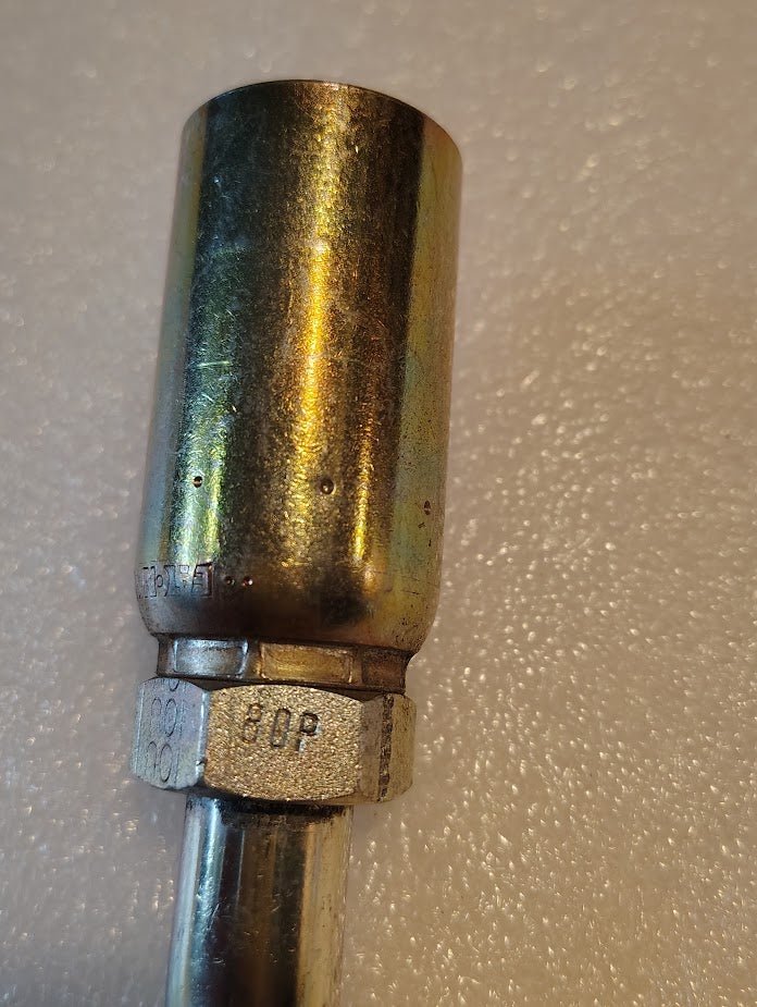 A close-up of the Weatherhead 10U-80P 5/8 Hose x 5/8 BSPP Female Fitting (CS46-10X10) shows its cylindrical body, hexagonal nut, and stamped markings on a textured, light surface—compatible with hydraulic hose fittings and 100R1 hoses.