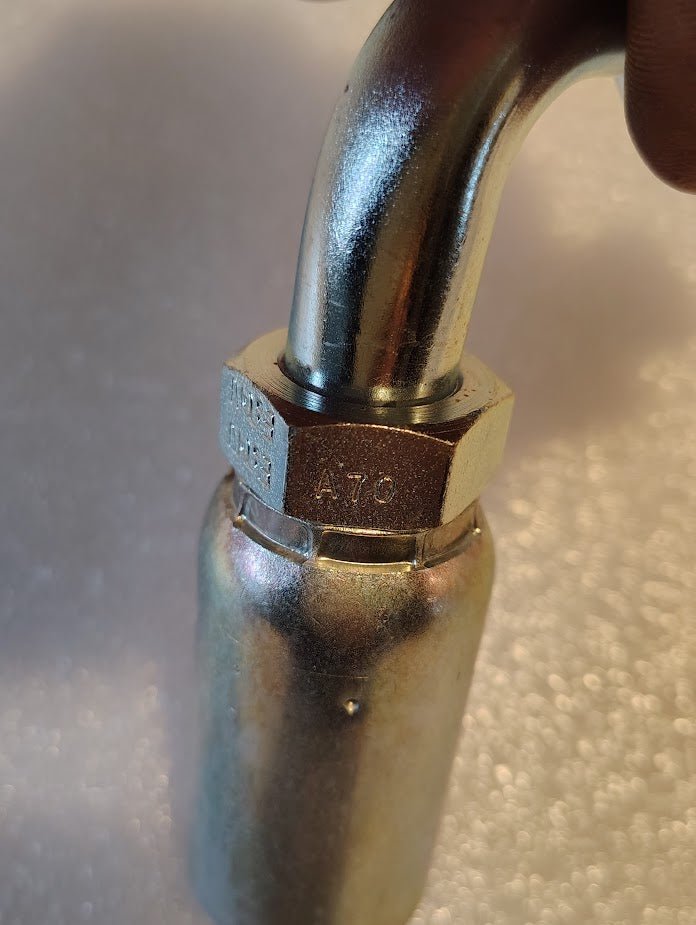 A close-up of a Weatherhead 10U-A70 (5/8 Hose x 5/8 ORF Female Fitting, CS33-10X10) with hex nut, attached to a curved pipe on textured white surface—ideal for hydraulic hose assemblies with 100R2 hoses.