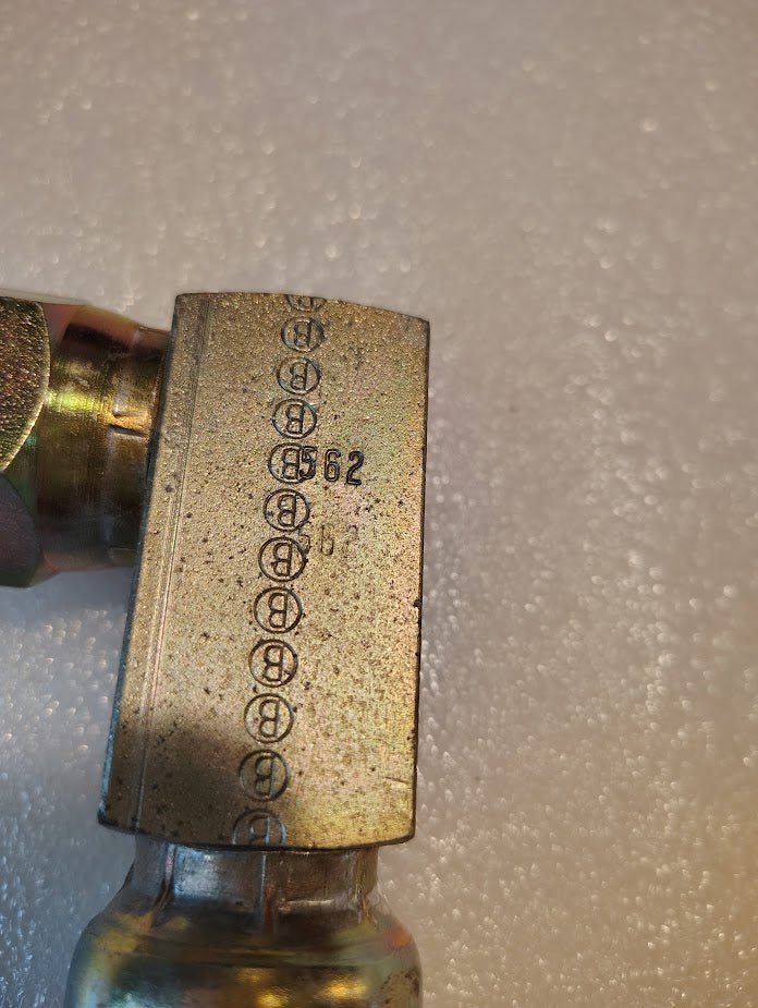 A close-up of a 12U-562 Weatherhead 3/4 Hose x 3/4 JIC Swivel Fitting - 90 Elbow shows engraved B symbols and the number 62 on its metal surface, with a plain, blurred background highlighting fine details.