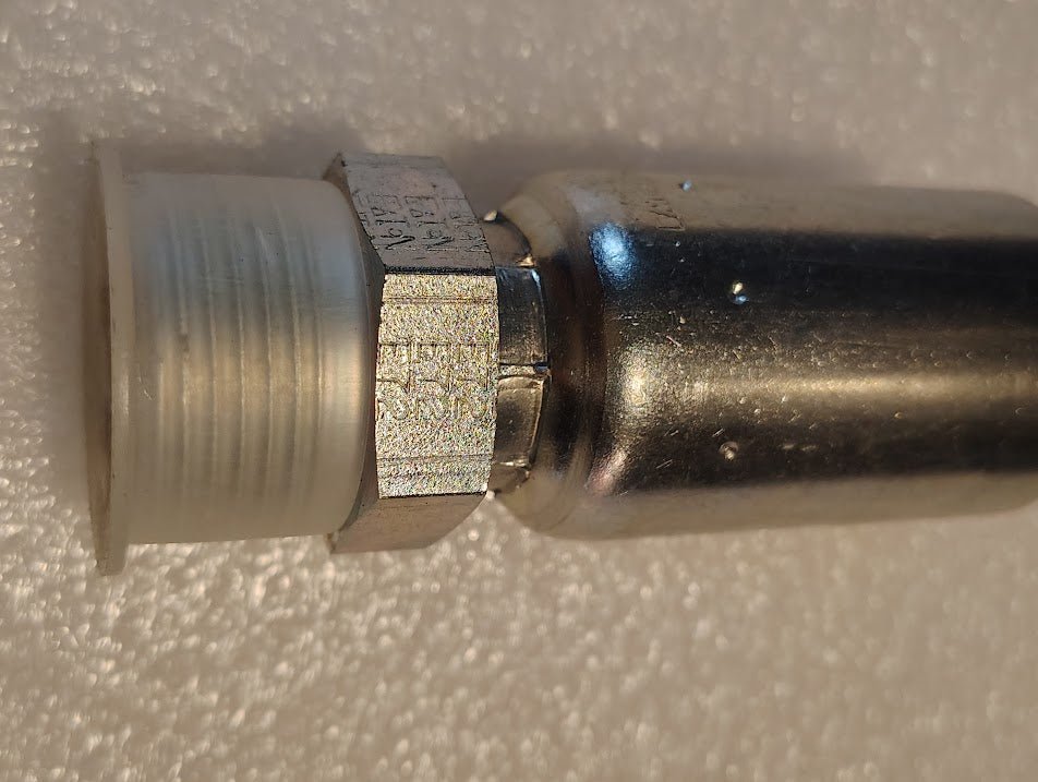 Close-up of a Weatherhead 12U-E72 (CS29-12X12), a 3/4 hose x 3/4 ORF male metal fitting with hex nut and white threaded connector, shown on a textured white surface.