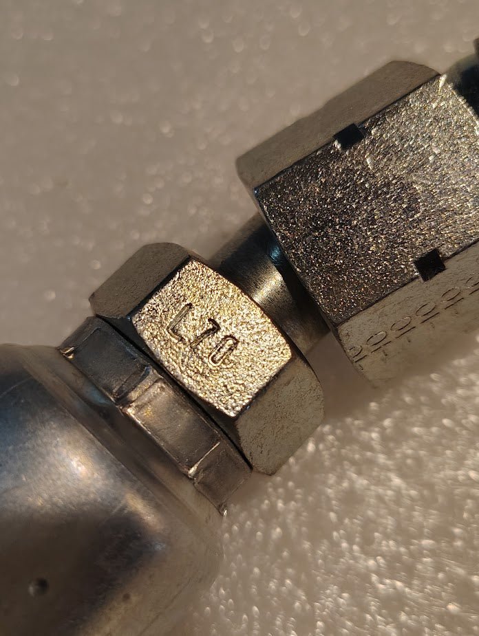 Close-up of a metal hex nut labeled L70 attached to a fitting, likely part of the Weatherhead 12U-L70 3/4 Hose x 5/8 ORF Female Fitting (CS31-12X10), set against a textured white background with shiny, detailed metallic surfaces.