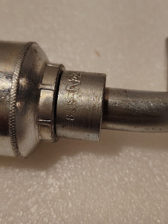 Close-up of product 43008U-668: a cylindrical metal object with a curved pipe attached. The worn metal shows partially visible engraved text 19-68 against a textured, light-colored background.