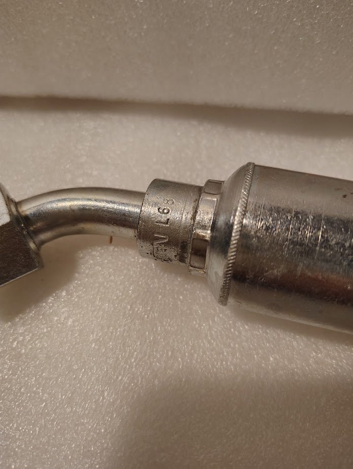 Close-up of a metal tool with a curved pipe attached to a Weatherhead 43008U-L68 (1/2 Hose x 1/2 ORFS Female Fitting, GW31-8x8), engraved with text and numbers, resting on a textured white surface.