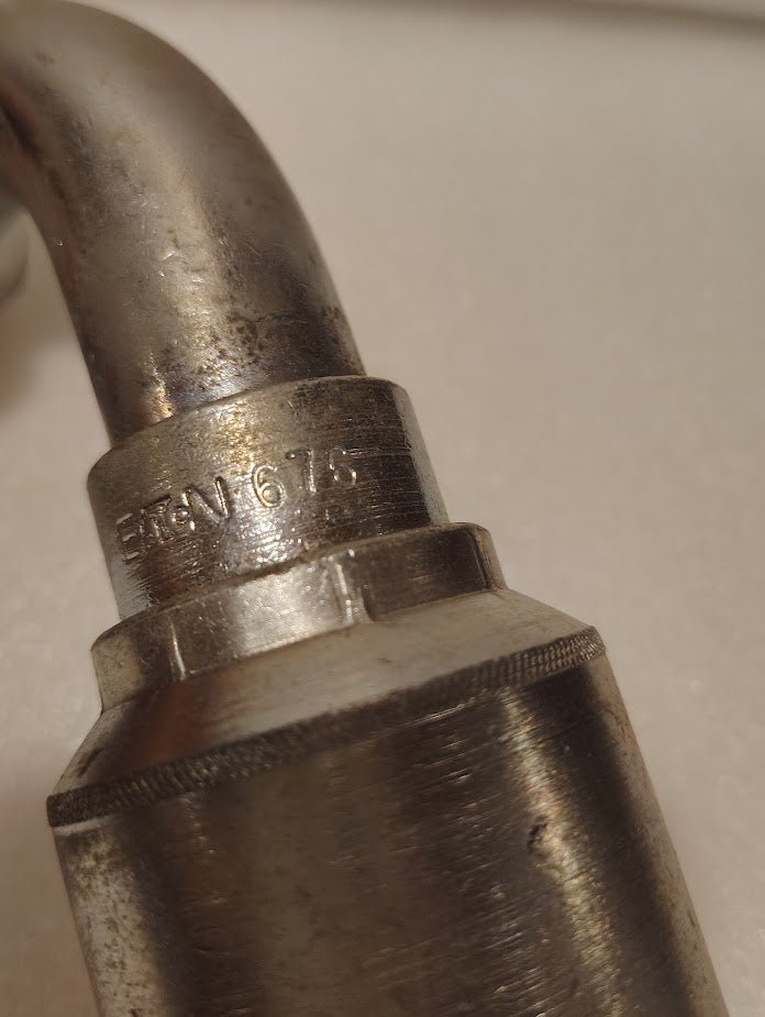 Close-up of a metallic pipe fitting engraved ETCN 674, similar to the 43016U-676 | 1 Hose x 1 JIC/SAE Swivel Fitting - 90 bend (GW26-16x16), with a slightly worn, curved female swivel at the top.