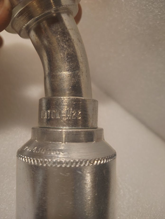 Close-up of a 43016U-H23 | 1 Hose x 1 Code 61 Flange Fitting - 30° bend (GW49-16x16) with hydraulic hose fittings and stamped A100122 on the connector. Out-of-focus white textured surface in the background.