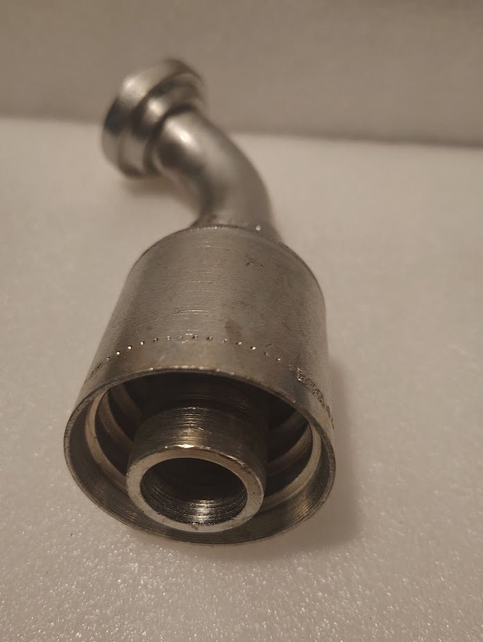 A close-up of the 43016U-H53 | 1 Hose x 1 Code 61 Flange Fitting with a 60° bend (GW51-16x16), metallic and threaded like hydraulic hose fittings, resting on a white textured surface.