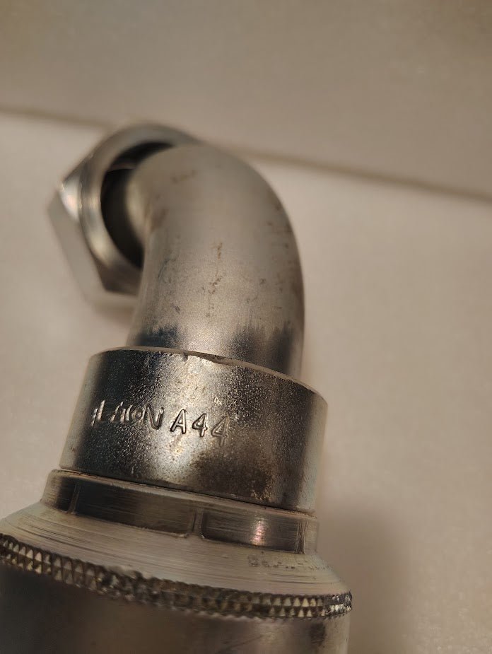 Close-up of a metallic, curved 43024U-A44 fitting (1 1/2 Hose x 1 1/2 ORFS Swivel, 90° short bend, GW32-24x24) with visible threading and engraved text, set against a plain light background.