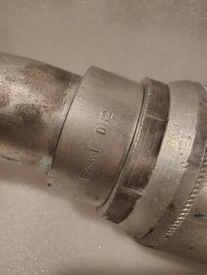 Close-up of a metal pipe fitting showing EATON and D82 engravings, likely the 43032U-D62 | 2 Hose x 2 Code 62 Flange Fitting - 45 bend (GW61-32x32), against a light background. The fitting displays visible signs of wear.