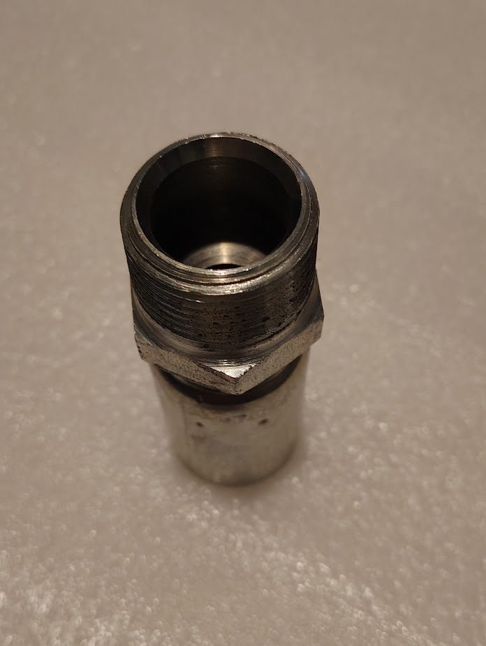 Close-up of a 12U-116 Weatherhead 3/4 Hose x 1 NPTF Male Fitting, featuring a cylindrical, shiny metal finish with thread on one end and some wear marks, standing upright on a textured surface.