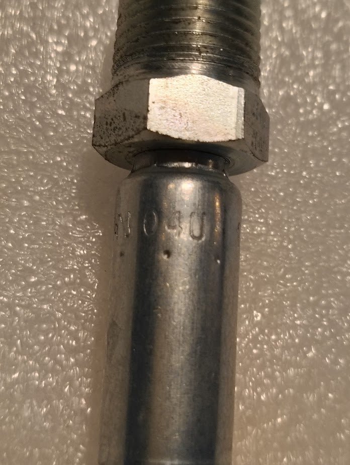 Close-up of a 04U-108 Weatherhead 1/4 Hose x 1/2 NPTF Male Fitting attached to a cylindrical pipe. Threading is visible on one end, with engraved numbers and minor wear on the metal surface, set against a textured, light-colored background.