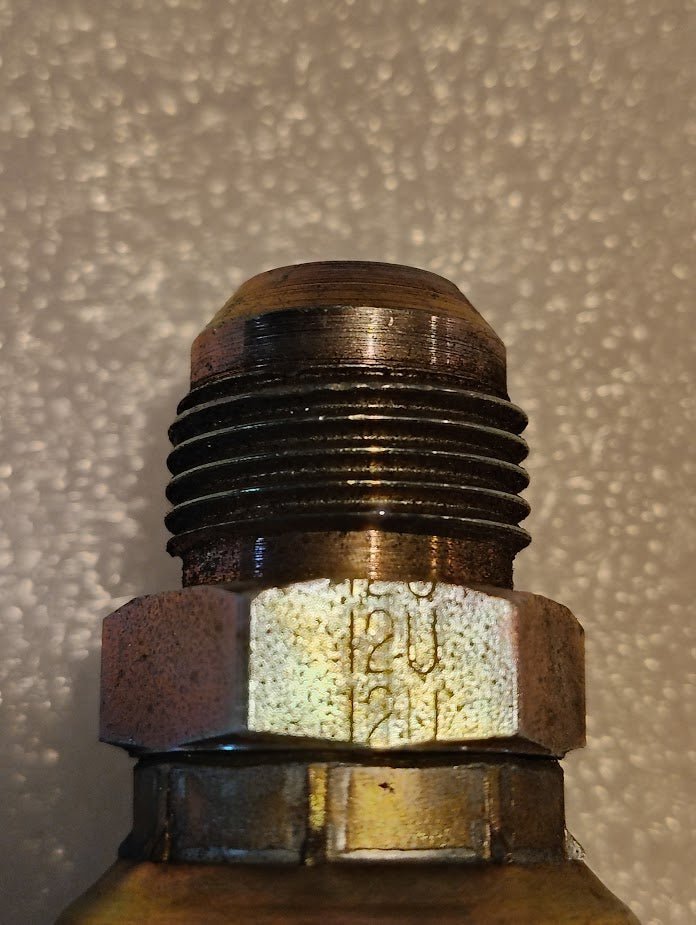 Close-up of the 12U-510 Weatherhead fitting—a straight solid bolt with a threaded section and hexagonal base against a textured, light backdrop. It resembles Parker fittings, with dark grooves, metallic shine, and visible markings on the hexagonal part.