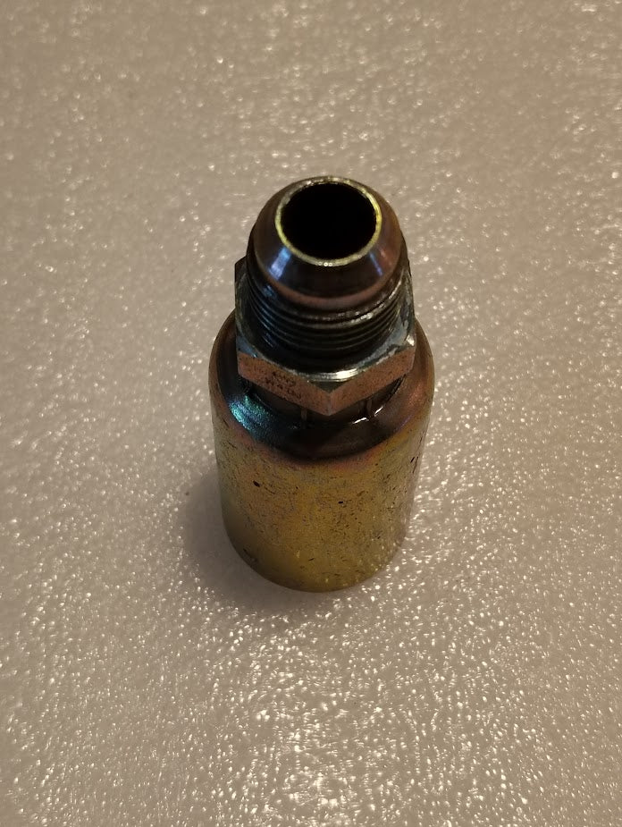 A metallic, cylindrical Weatherhead 12U-510 3/4 Hose x 5/8 JIC Male Fitting stands upright on a textured, light-colored surface. Slightly worn with discolored and shiny areas, it hints at past use in hydraulic hose systems.