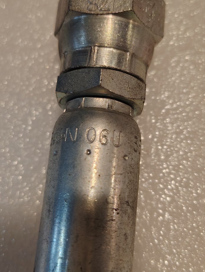 A close-up of the 06U-358 Weatherhead 3/8 Hose x 1/2 BSPP Female Fitting - Straight Swivel with alphanumeric markings lies on a textured white surface. The metallic fitting features a hexagonal coupling and shows slight wear.