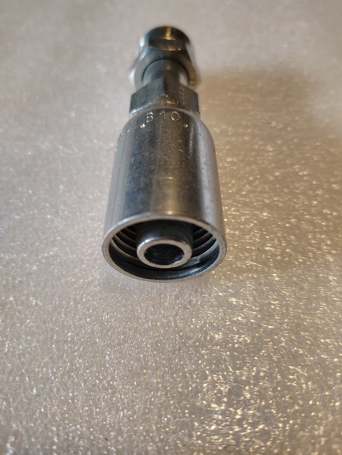 A close-up of a metallic part resembling hydraulic hose fittings lies on a textured surface. This 08U-B10 Weatherhead 1/2 Hose x 5/8 Inverted Male Fitting - Straight Swivel has a threaded interior and tapered end, casting a shadow beneath it.