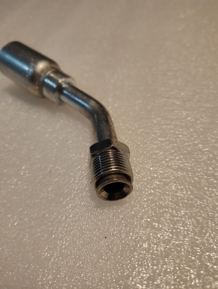 A metal pipe fitting with a threaded end rests on a textured white surface. It resembles the 08U-B48, a Weatherhead 1/2 Hose x 1/2 Inverted Male Fitting with a 45-degree swivel, and appears shiny, suggesting it might be stainless steel or similar material.