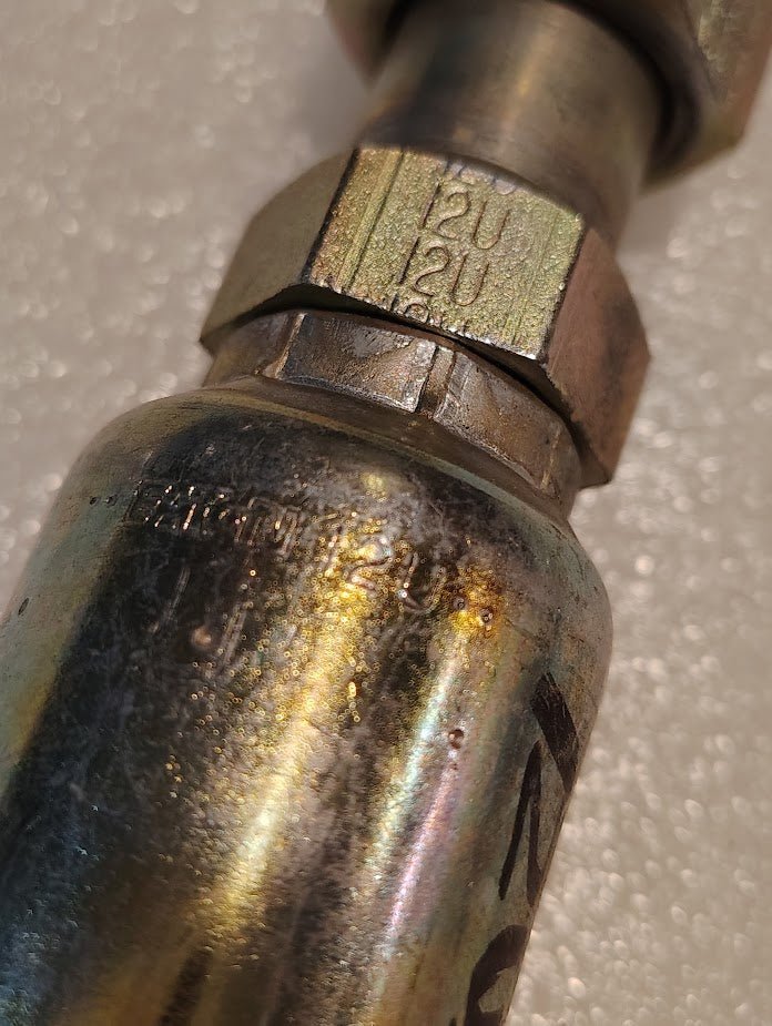 A detailed view of a slightly worn 12U-S70 Weatherhead fitting with visible threads and 12U markings shows iridescent hues. Essential for 100R1 hydraulic hoses, it is set against a soft, light backdrop.