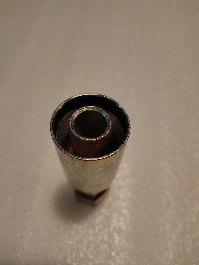 A metallic cylindrical object with an inner threaded section stands upright on a textured white surface. Resembling a hydraulic hose fitting, this component is likely the 12U-S70 Weatherhead 3/4 Hose x 5/8 ORF Female Fitting - Straight Swivel, used in plumbing or machinery.
