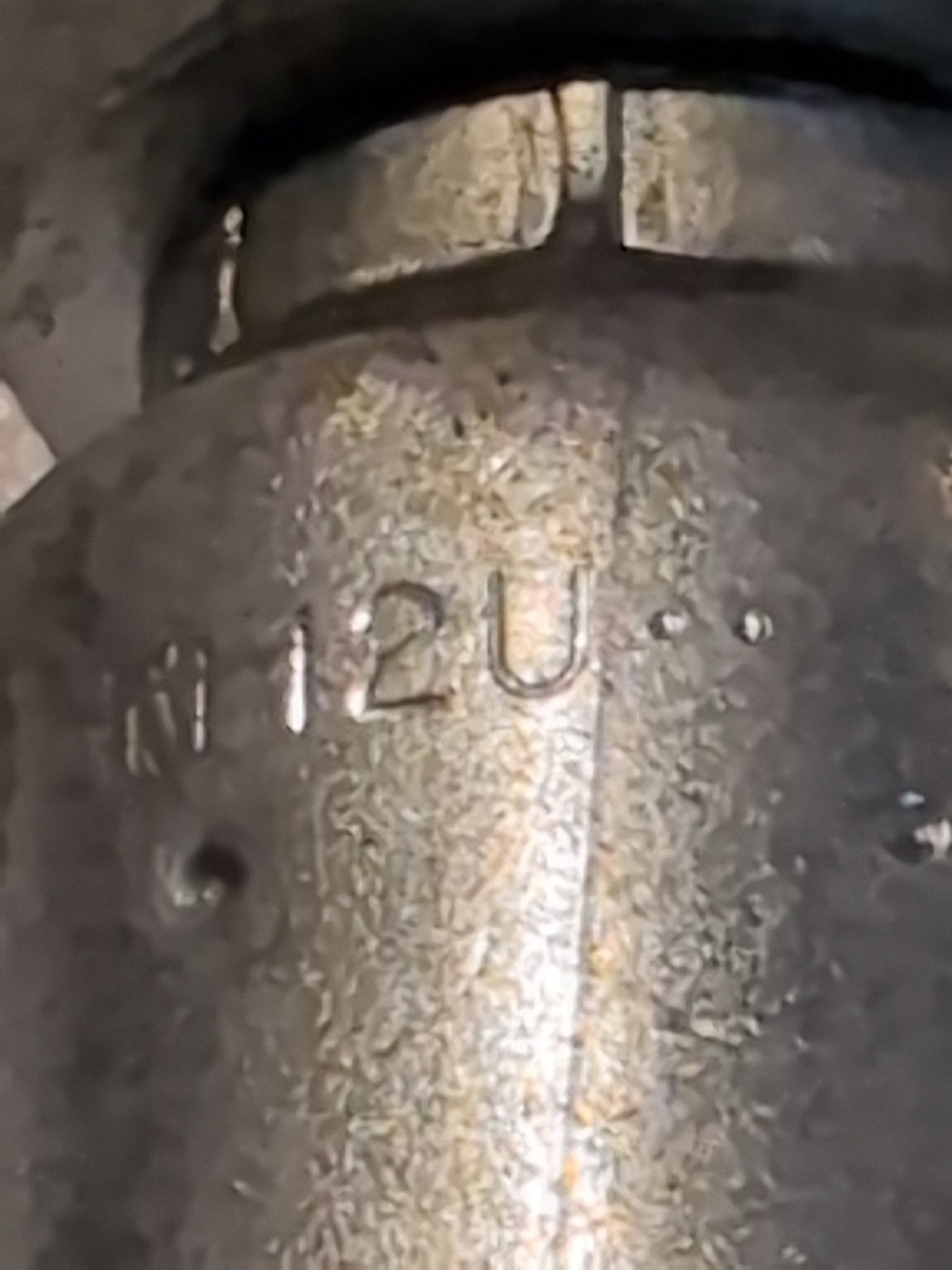 A close-up of a textured metal surface, similar to the Weatherhead 12U-S76 fitting, engraved with 12U and other indistinct marks. The slightly tarnished look suggests wear and age like that found on well-used hydraulic hose fittings.