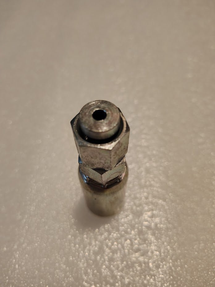 A close-up of a 06U-S64 Weatherhead fitting shows the metallic hexagonal design with a narrow opening, positioned vertically on a textured white surface. The shiny, reflective finish mirrors the quality associated with Weatherhead fittings.