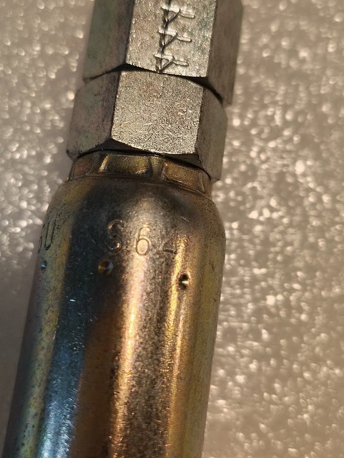 Close-up of a metallic fitting with a hexagonal connector, compatible with 100R1 hydraulic hose fittings. It features engraved S64 markings and is set against a textured, bumpy white background. This is the Weatherhead 06U-S64: a 3/8 hose x 1/4 ORF female fitting - straight swivel.
