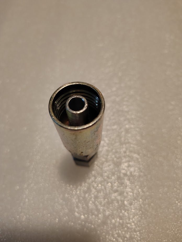 A close-up image shows a Weatherhead 06U-S64 fitting—metallic, cylindrical, and threaded inside. Its positioned upright on a textured white surface.