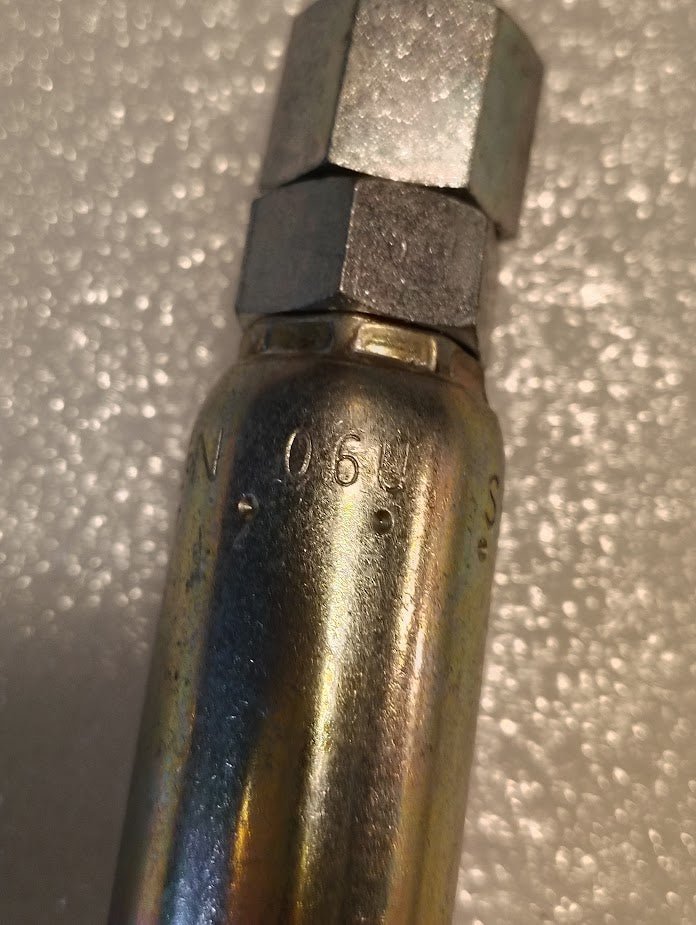 The image is a close-up of the 06U-S64 Weatherhead fitting, featuring engraved 0.6 U.S. on a metallic cylindrical form with a hexagonal top akin to hydraulic hose fittings. Its slightly reflective, textured surface stands out against the blurred background.