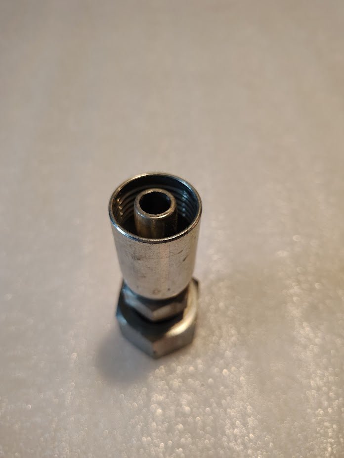 A close-up of the 08U-S72 Weatherhead fitting, a shiny silver coaxial connector resembling Parker fittings with a hexagonal nut, stands upright on a light textured surface.