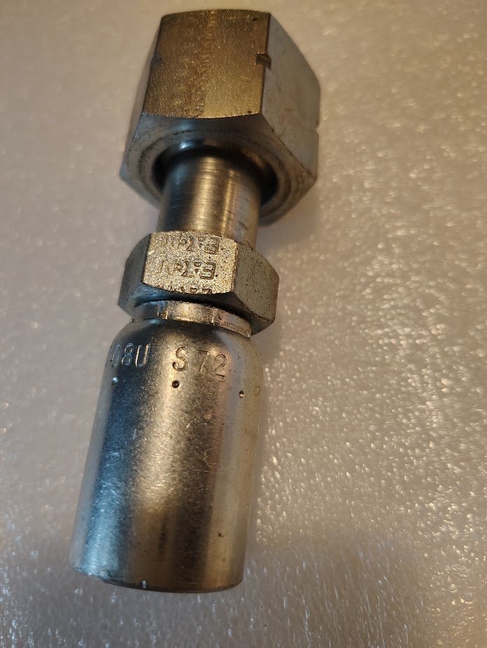 A close-up of a metallic hydraulic hose fitting sits on a textured white surface. This high-quality 08U-S72 Weatherhead fitting, resembling others with components like a hexagonal nut and cylindrical parts, includes visible inscriptions and markings.