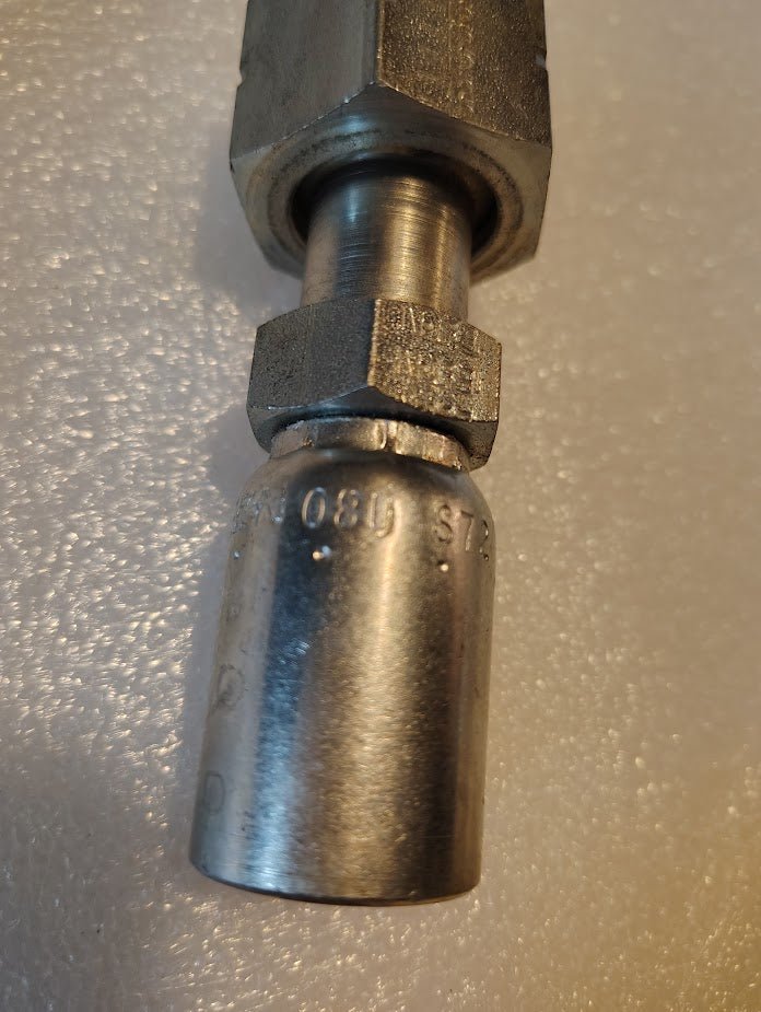 A close-up of a silver, cylindrical 08U-S72 Weatherhead 1/2 Hose x 3/4 ORF Female Fitting with threaded connectors on a textured white surface. Engraved details indicate 100R1 compatibility, ensuring seamless alignment for precise connections.