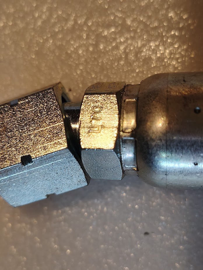 Close-up of a metal mechanical component featuring a hexagonal nut with engraved markings, connected to a textured metal fitting: the 10U-L70 Weatherhead 5/8 Hose x 5/8 ORF Female Fitting - 45 Swivel. The background is a plain, light-colored surface.