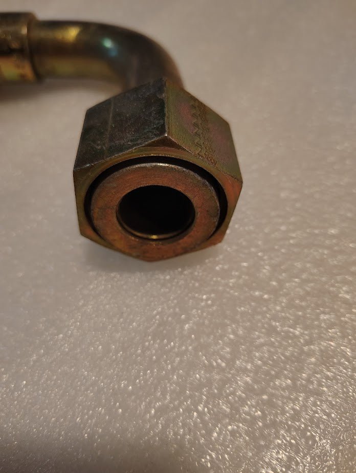 Close-up of the Weatherhead 16U-A76 1 Hose x 1 ORF Female Fitting. It features a hexagonal nut with a dark circular opening and has a 90-degree elbow joint extending to the left, resting on a textured white surface.