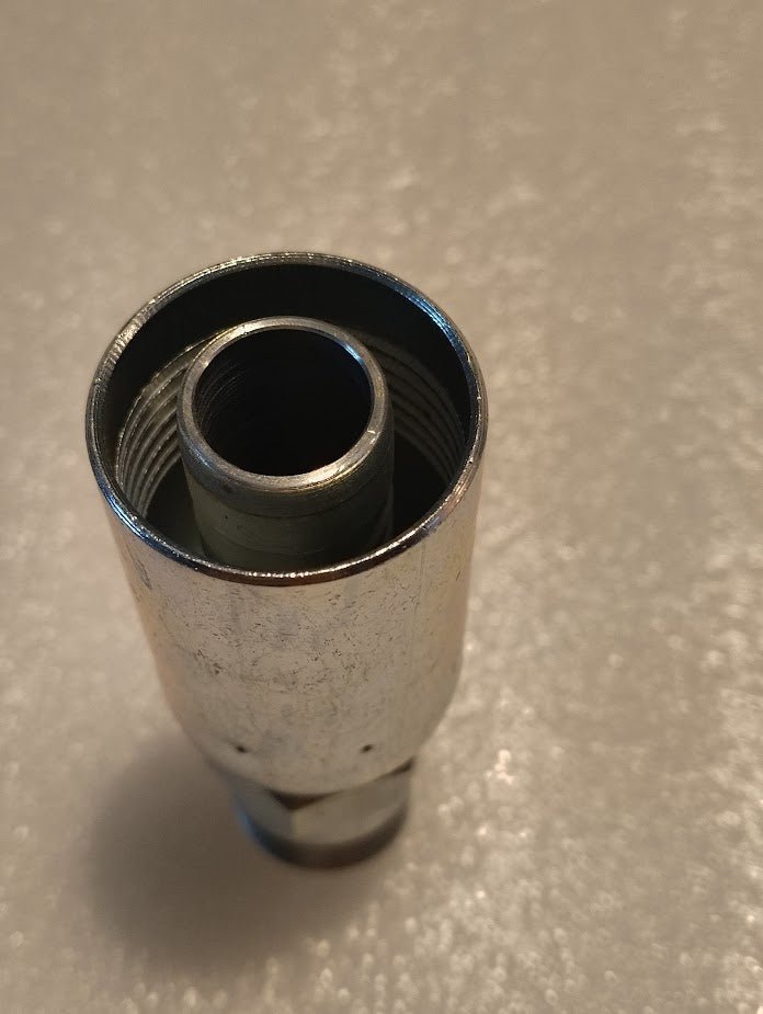 A close-up of a Weatherhead 3/4 Hose x 3/4 BSPT Male Fitting - Straight Solid (12U-162) shows its metallic, cylindrical form with internal threading, standing upright on a textured surface. The reflective object is designed for efficient connecting or fastening through its hollow center.