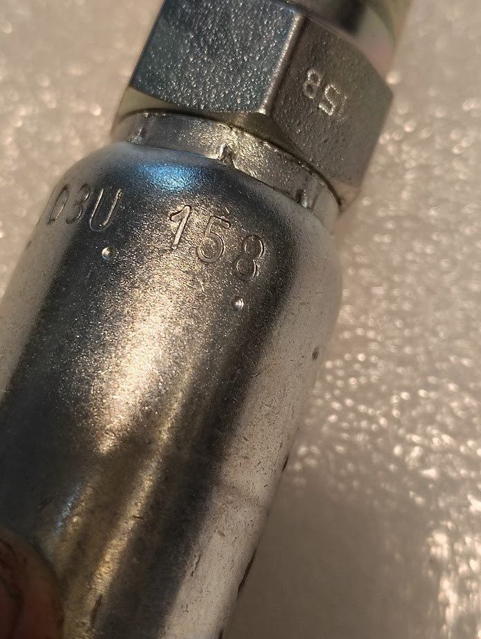 Close-up of a metallic component labeled 08U-158, akin to Weatherhead 1/2 Hose x 1/2 BSPT Male Fitting - Straight Solid, set against a textured white surface. The shiny finish and reflections highlight its intricate design.